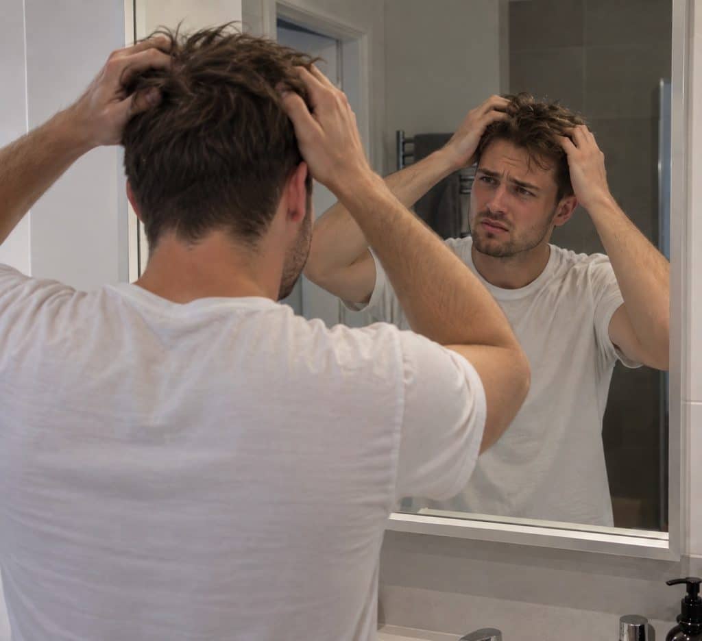 A man with his hands in his hair, looking at his reflection in a mirror